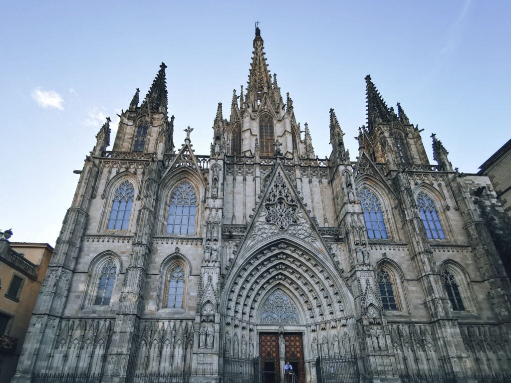 A large Gothic cathedral with pointed spires, ornate stone carvings, and a central arched entrance under a clear blue sky.