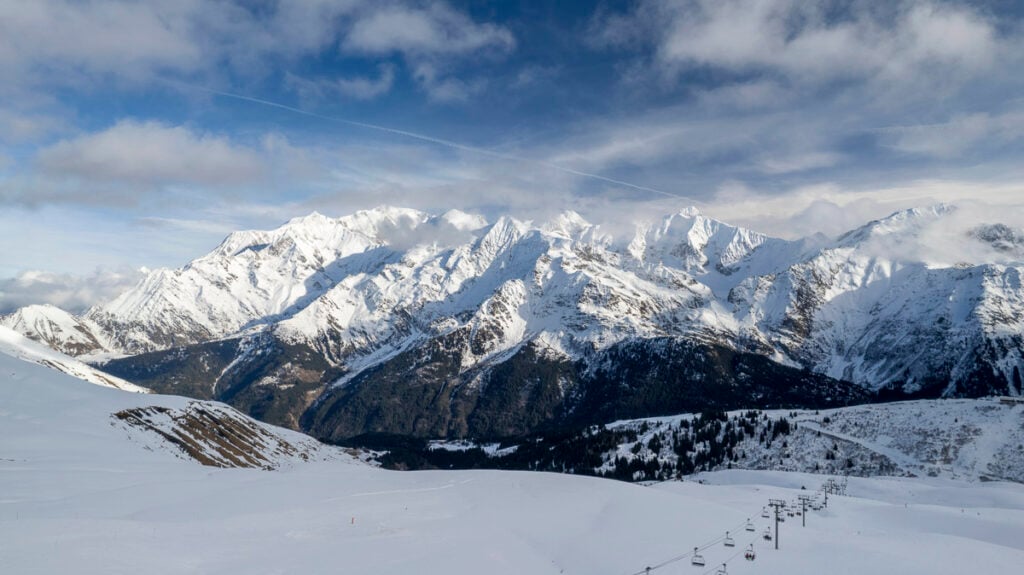 Snow-covered mountains under a partly cloudy sky with a ski lift visible in the lower right corner.