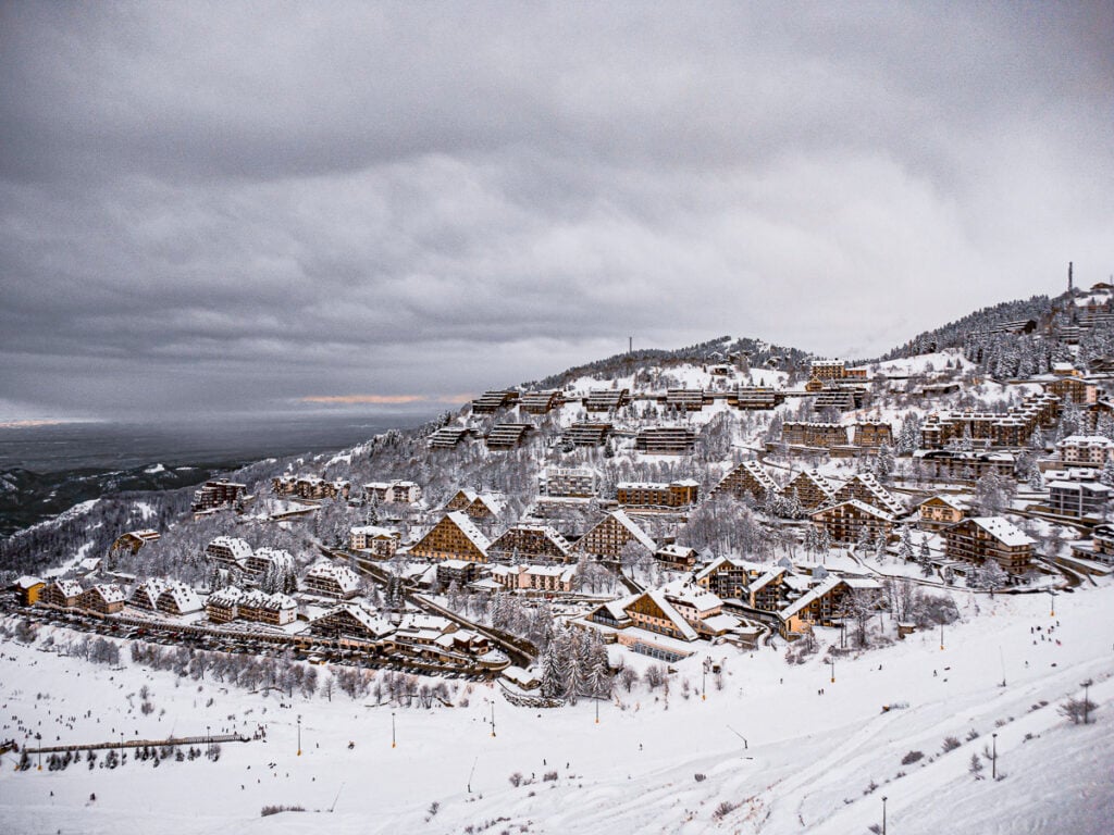 Snow-covered mountain village with multiple chalet-style buildings under a cloudy sky, surrounded by snowy slopes and trees.