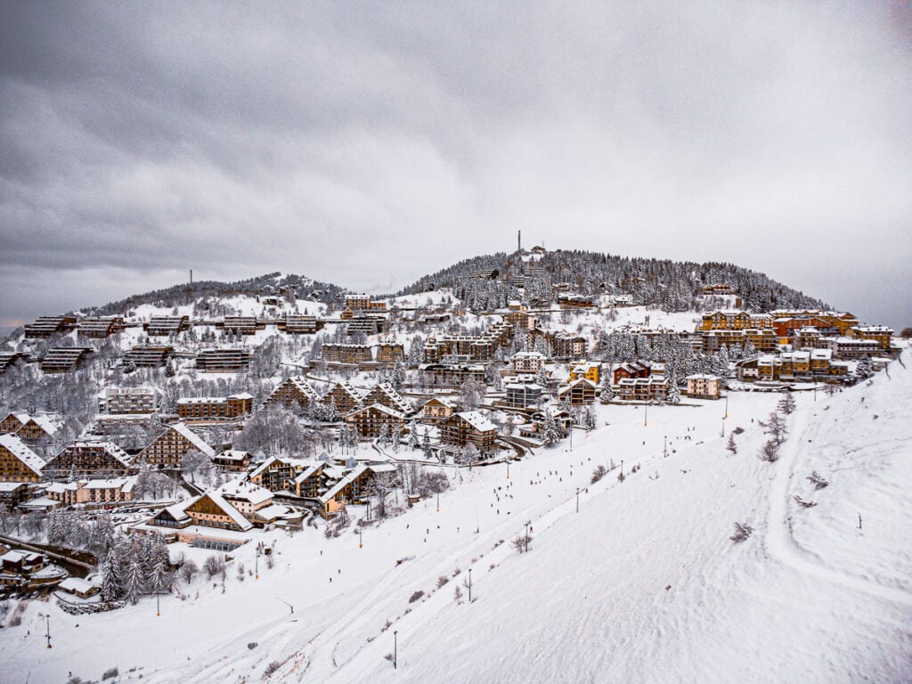 Snow-covered buildings and trees on a hillside under a cloudy sky, with people skiing or walking on the slopes in the foreground.