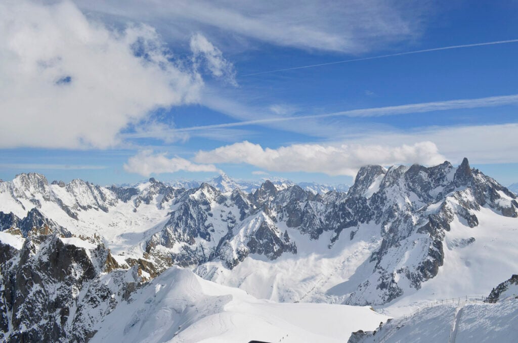 Snow-covered mountain peaks under a blue sky with scattered clouds and visible contrails.