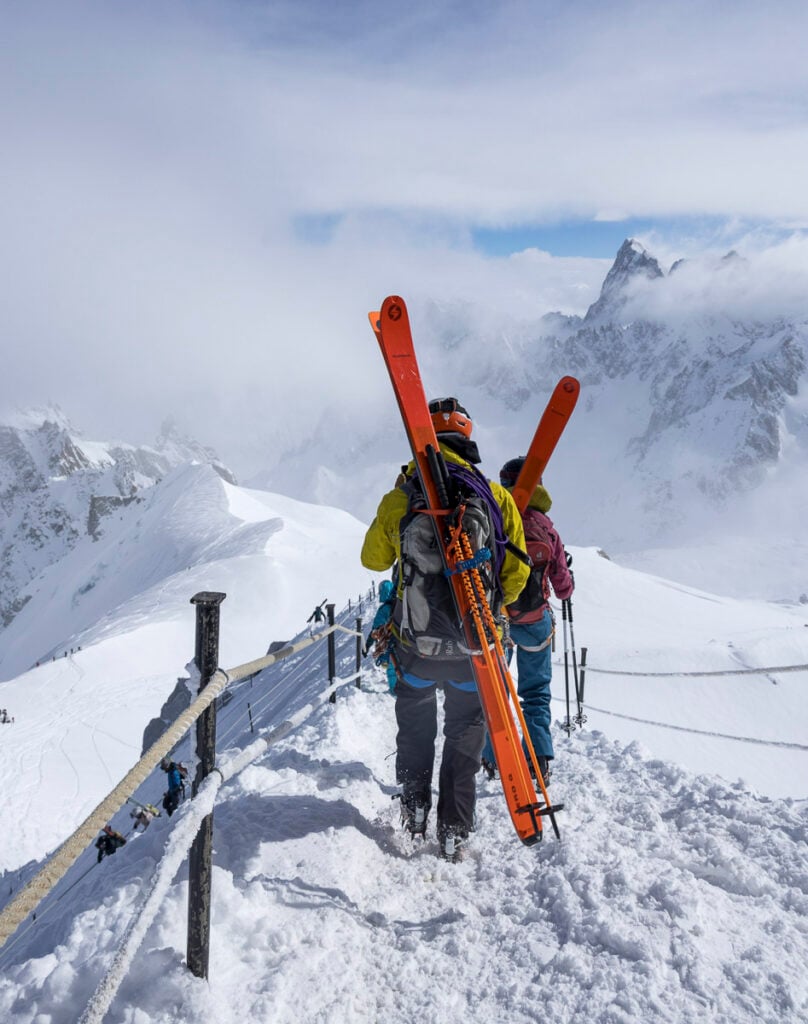 Two skiers carrying orange skis walk along a snowy mountain ridge, surrounded by snow-covered peaks under a cloudy sky.