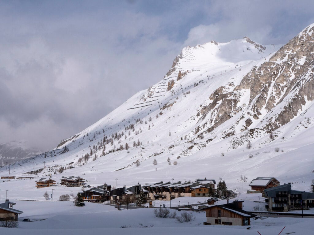 Snow-covered mountain with scattered trees rises behind a small alpine village of wooden buildings under a cloudy sky.