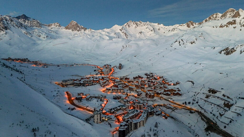 Aerial view of a snow-covered mountain resort town at dusk, with illuminated streets winding between buildings and surrounding peaks visible in the background.