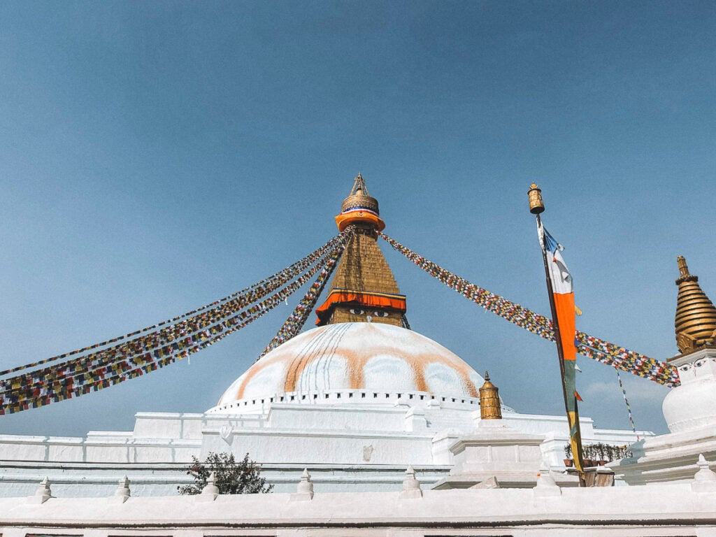 A large white stupa with a golden spire and colorful prayer flags against a clear blue sky.