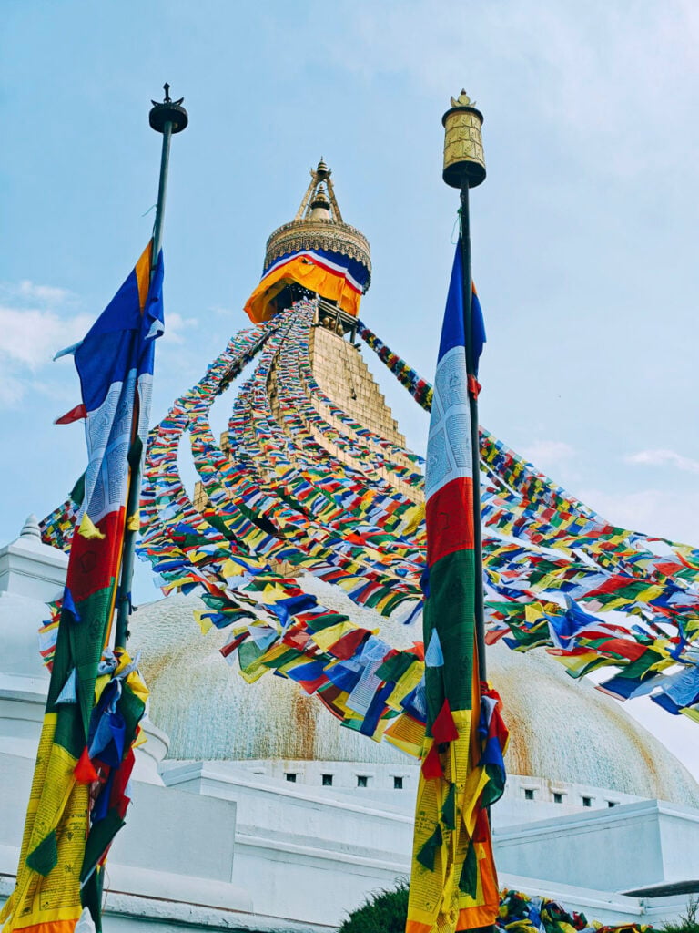 A large stupa with rows of colorful prayer flags extending from its spire, flanked by two flagpoles, under a clear sky.