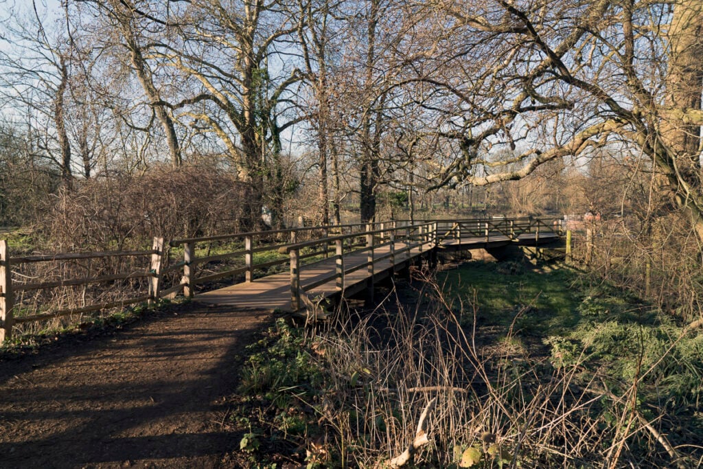 Wooden pedestrian bridge crosses over a small stream in a park surrounded by leafless trees and undergrowth on a sunny day.