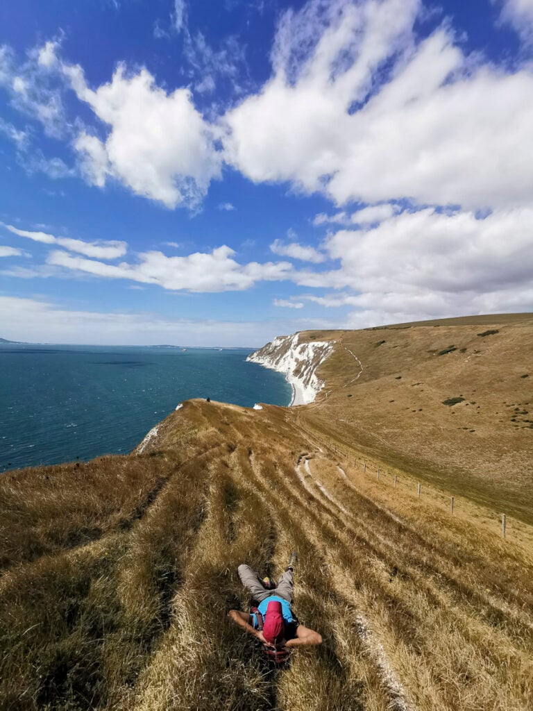 A person lies on dry grass overlooking white cliffs and the sea under a partly cloudy sky.