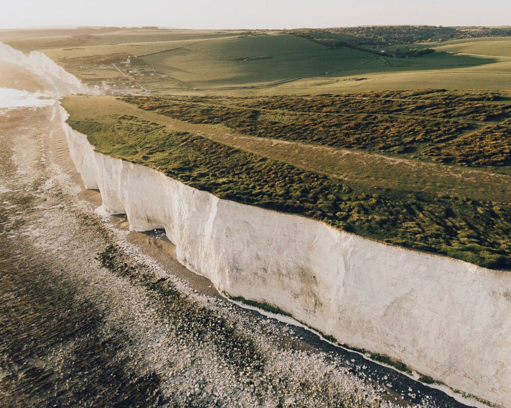Aerial view of tall white chalk cliffs meeting the sea, with green grassy fields on top and a rocky shoreline below.