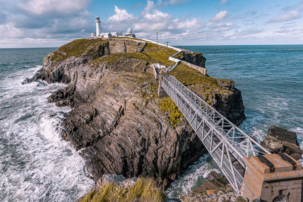 A metal footbridge leads to a rocky island with a lighthouse and buildings, surrounded by ocean waves under a partly cloudy sky.