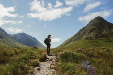 A person wearing a backpack stands on a rocky path between grassy hills under a blue sky with scattered clouds.