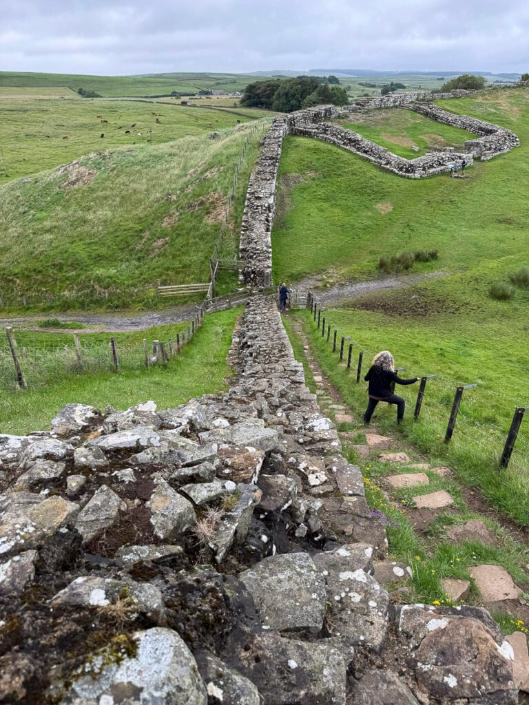 A person climbs stone steps along Hadrian’s Wall, which stretches across a grassy landscape under a cloudy sky.
