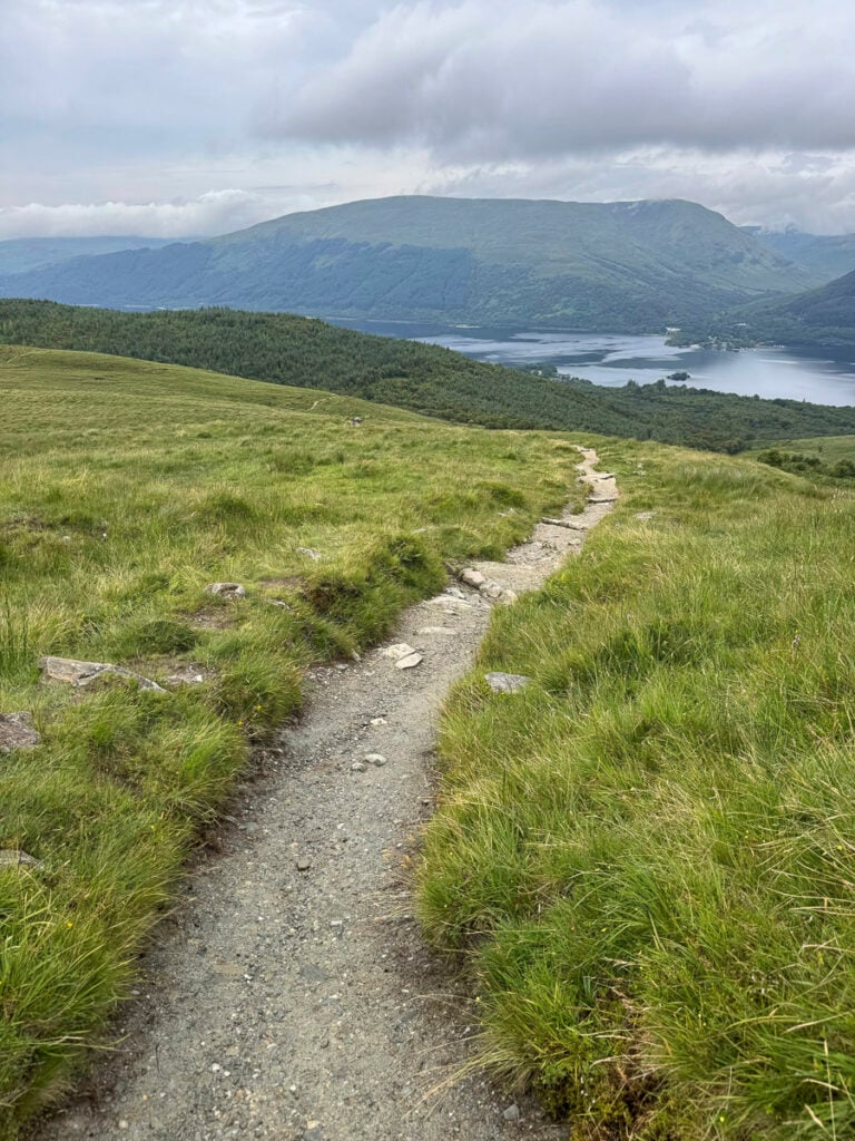 Narrow gravel path through grassy hills leading toward a lake and mountains under a cloudy sky.