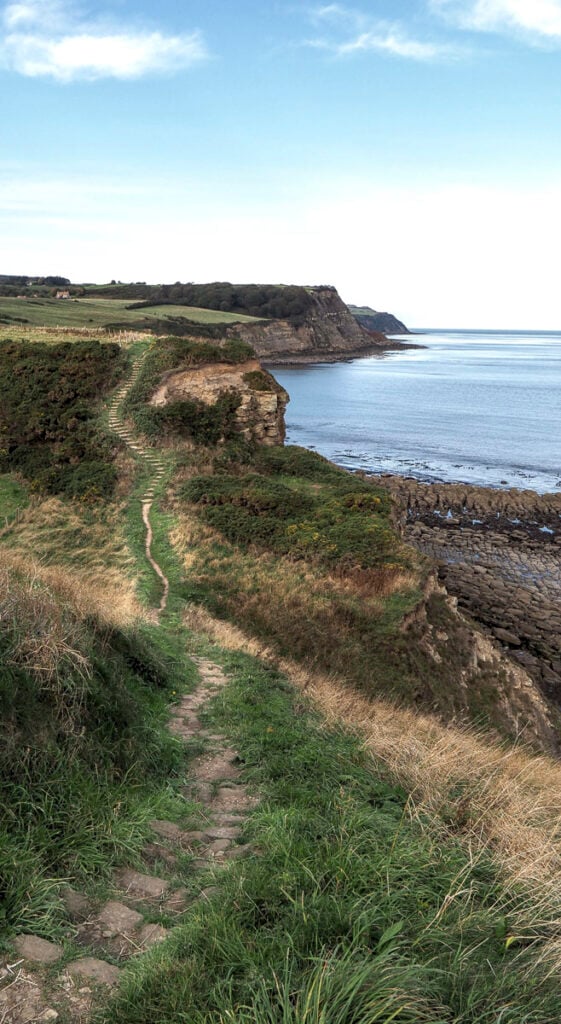 Narrow stone path winds along grassy coastal cliffs, overlooking calm blue sea and rocky shoreline under a partly cloudy sky.