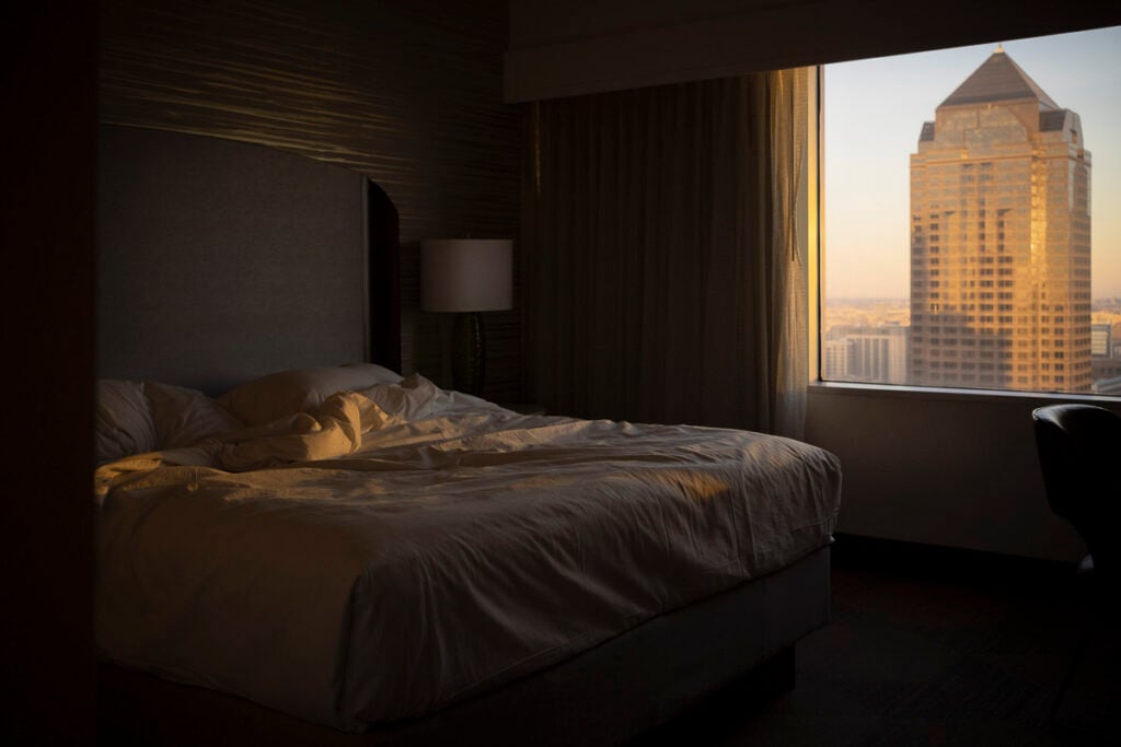 A hotel room with a made bed, illuminated by warm sunlight streaming through a window that overlooks a city skyscraper.
