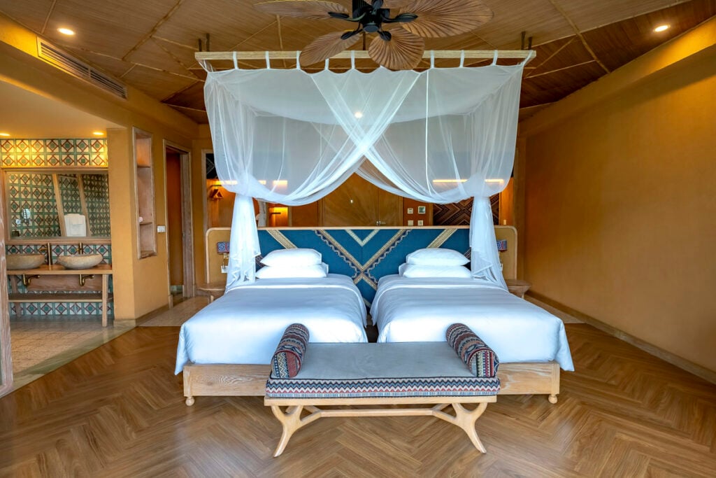 A hotel bedroom with two twin beds featuring white bedding, a draped mosquito net canopy, and a decorative bench at the foot of the beds.