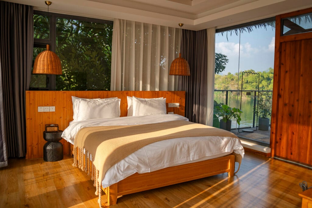A bedroom with a wooden bed frame, white bedding, and a beige throw blanket, set against a wooden accent wall with large windows overlooking a lake.