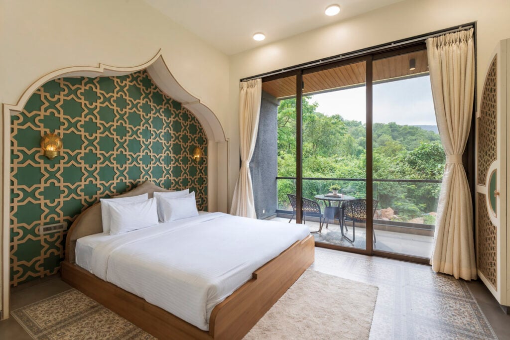 A hotel bedroom with a queen bed set against an arched green and gold patterned wall, with a glass door leading to a balcony overlooking a wooded area.
