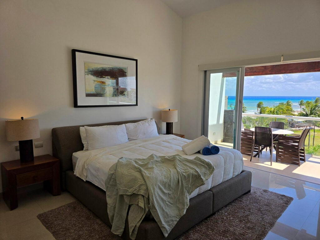 A hotel room with a queen bed, two nightstands, and a glass door opening onto a balcony with an ocean view.